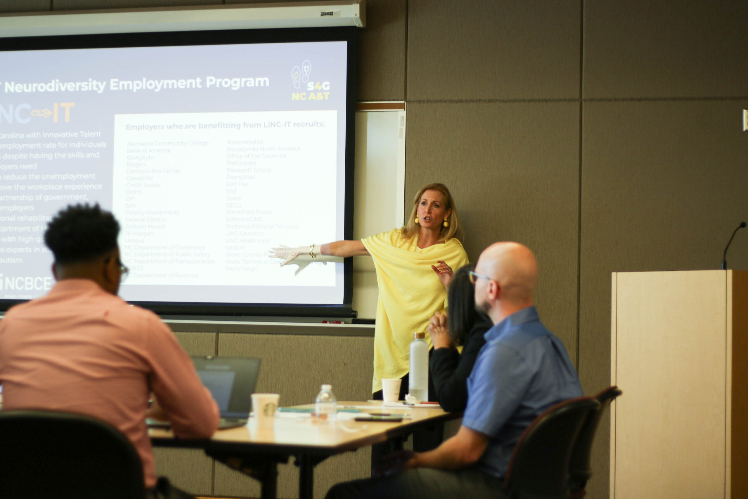 A woman in a yellow outfit is shown at the front of a room presenting on a neurodiversity employment program. Two people are shown at a table listening to her. One is typing on a computer.