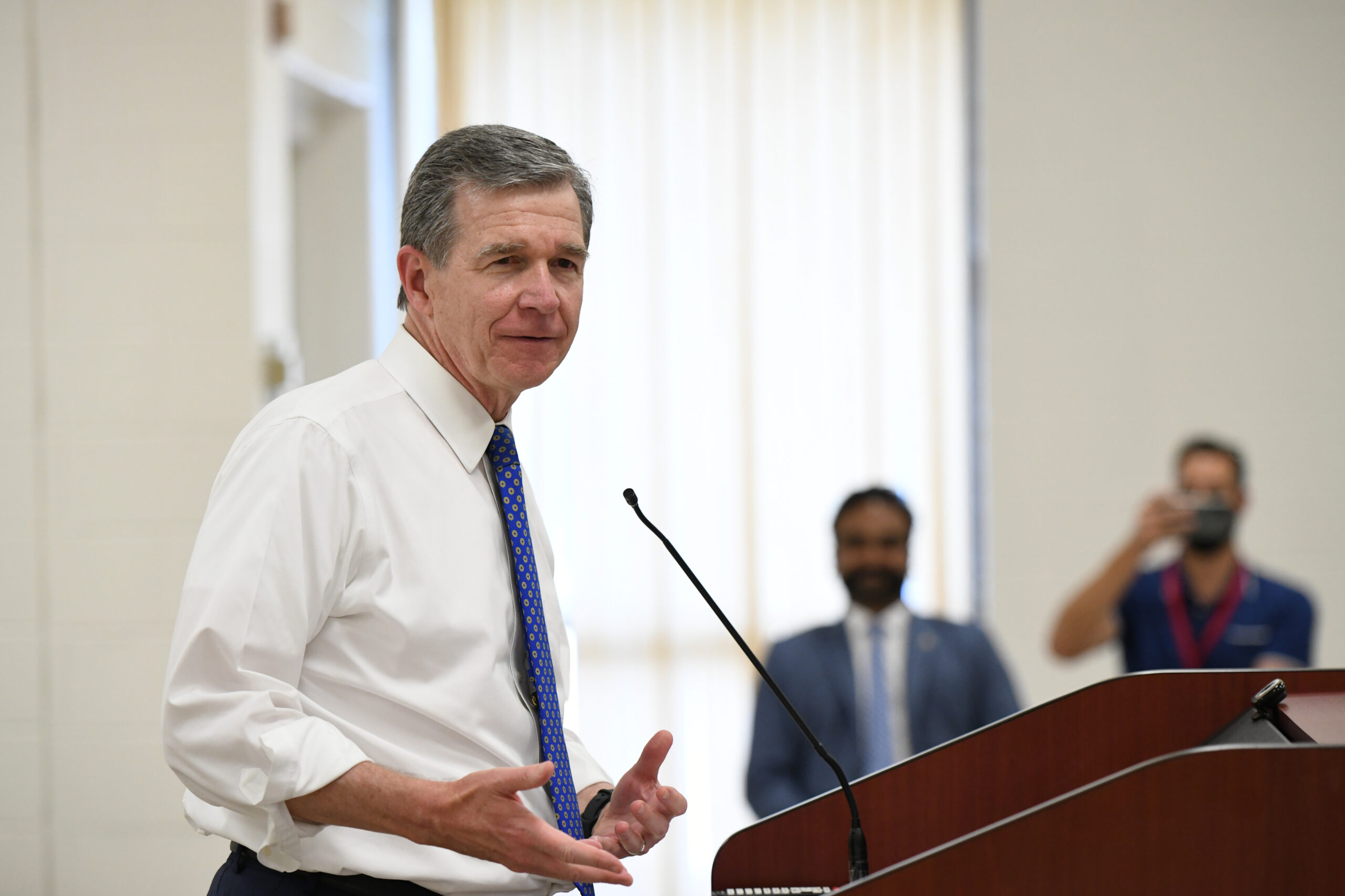 Governor Roy Cooper is shown speaking at a podium. Two people are in the background listening. One person is taking a photo.