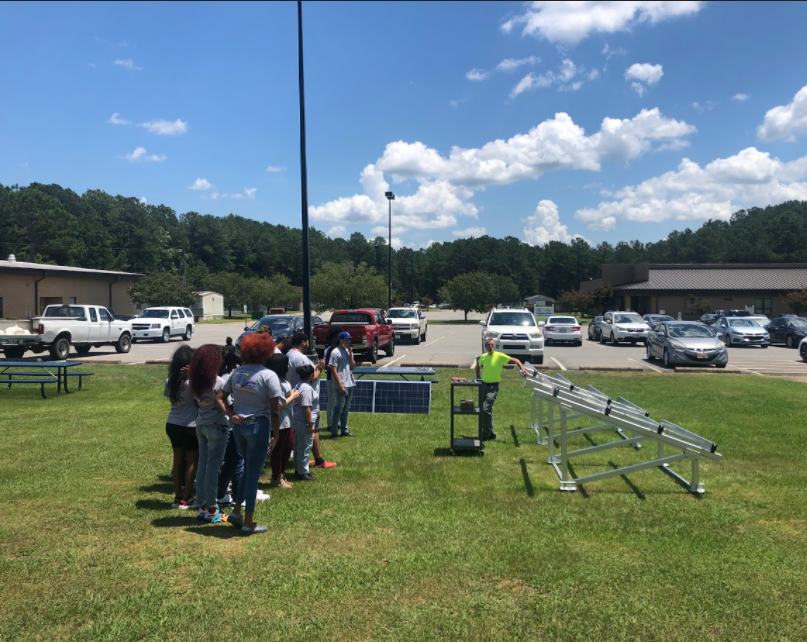 A group of students wearing gray t-shirts and jeans are listening to a man in a neon yellow shirt speak. He is leaning against a solar panel stand and has a cart near him. There is a solar panel past him and the students. They are all outside in the grass next to a parking lot.